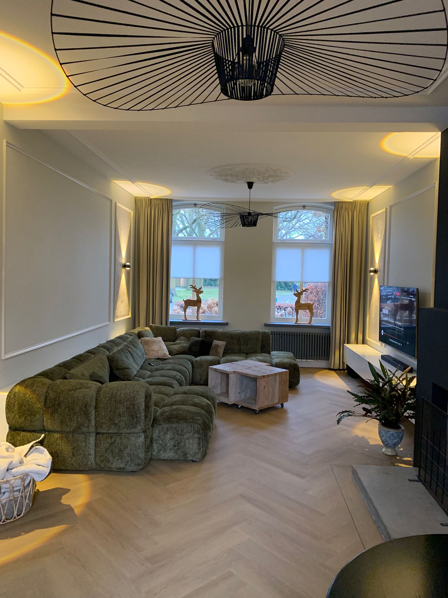 Lichte visgraat houten vloer in een stijlvolle woonkamer, gecombineerd met een groene hoekbank, warme verlichting en een moderne, sfeervolle inrichting.